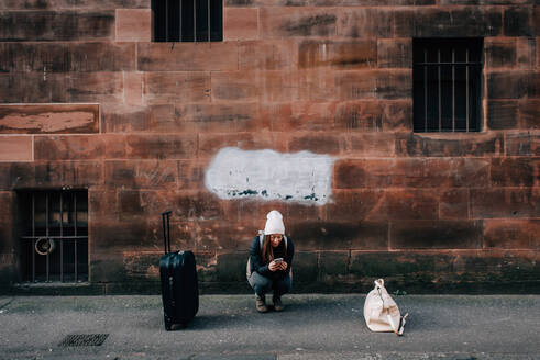 Frau, die mit Handy und Gepäck eine SMS auf dem Bordstein schreibt, Edinburgh, Schottland - CUF52493