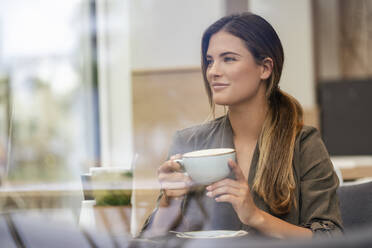 Junge Geschäftsfrau mit Kaffeetasse in einem Café, durch ein Fenster gesehen - DIGF07117