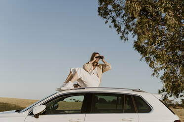 Weibliche Reisende schaut durch ein Fernglas auf einem weißen Autodach sitzend - ERRF01601