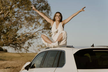 Smiling female traveller sitting on white car roof, raised arms - ERRF01606