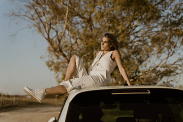 Female traveller sitting on white car roof - ERRF01607