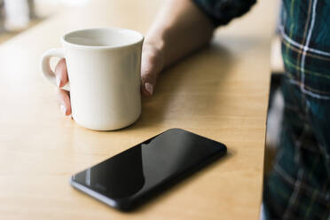 Hand of young woman holding cup of coffee next to smartphone - JOSF03318