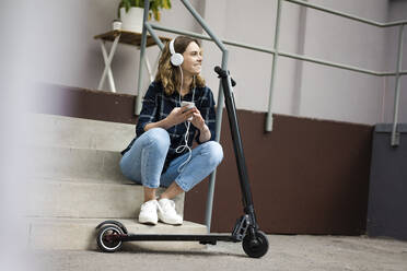 Young womanwith electric scooter, sitting on stairs, listening music - JOSF03319