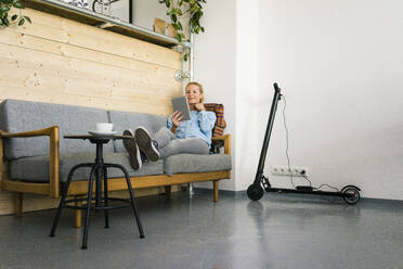 Junge Frau sitzt auf einer Couch in einem Café und benutzt ein digitales Tablet, während im Hintergrund ein E-Scooter aufgeladen wird - JOSF03338