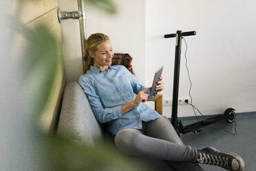 Junge Frau sitzt auf einer Couch in einem Café und benutzt ein digitales Tablet, während im Hintergrund ein E-Scooter aufgeladen wird - JOSF03339