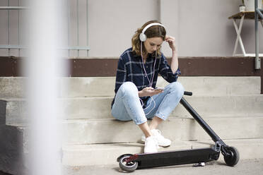 Junge Frau mit Elektroroller, sitzt auf der Treppe und hört Musik - JOSF03362