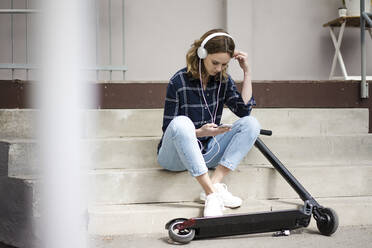 Young womanwith electric scooter, sitting on stairs, listening music - JOSF03362