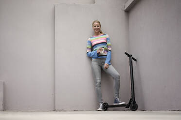 Young woman with electric scooter in a loft, holding cup of coffee - JOSF03371