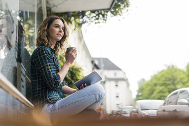 Junge Frau, die vor einem Café sitzt, ein digitales Tablet benutzt und Kaffee trinkt - JOSF03433