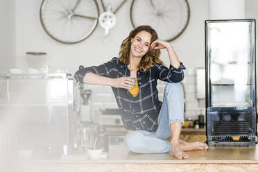 Young woman sitting barefoot on counter of her own coffee shop, drinking coffee - JOSF03438