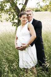 Pregnant bride with his husband holding baby belly on a meadow - HMEF00473