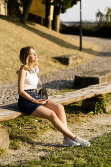 Lächelnde junge Frau mit Kopfhörern und Smartphone sitzt auf einer Bank und genießt das Sonnenlicht - GIOF06576