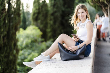 Portrait of smiling young woman with headphones and smartphone sitting on a wall - GIOF06577
