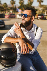 Portrait of bearded motorcyclist with mirrored sunglasses leaning on his helmet smoking - LJF00293