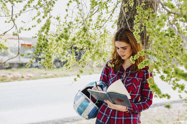 Redheaded woman with motorcycle helmet leaning against tree taking notes - LJF00352