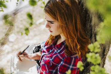 Pensive redheaded woman leaning against tree trunk taking notes - LJF00355