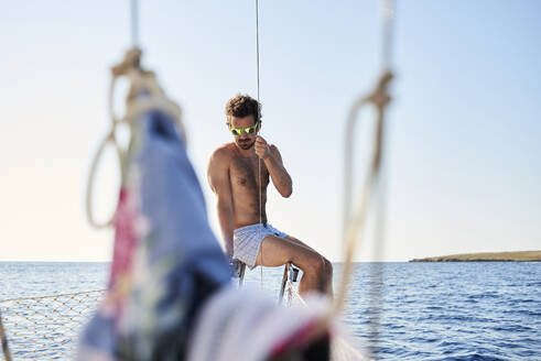Mann in Boxershorts auf einem Segelboot sitzend, Menorca, Spanien - IGGF01254