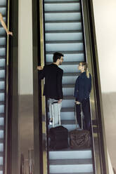 Zwei junge Geschäftspartner mit Gepäck im Gespräch auf einer Rolltreppe - JSRF00377