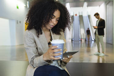 Young businesswoman with takeaway coffee and smartphone in a foyer - JSRF00405
