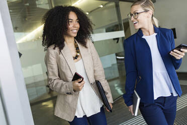 Two happy young businesswomen walking and talking in a hallway - JSRF00422