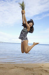 Mixed race teenager jumping and holding pineapple on beach - BLEF08341