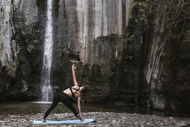Woman practising yoga at waterfall, triangle pose - LJF00363