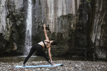 Woman practising yoga at waterfall, triangle pose - LJF00364