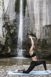 Woman practising yoga at waterfall, warrior pose - LJF00368