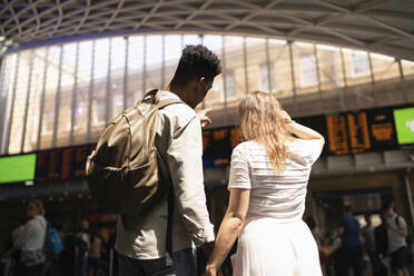 Rückenansicht eines jungen Paares, das die Anzeigetafel eines Bahnhofs betrachtet, London, UK - FBAF00853