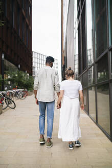 Rückenansicht eines jungen Paares, das Hand in Hand auf dem Bürgersteig geht - FBAF00874