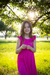 Porträt eines kleinen Mädchens in einem leuchtend rosa Kleid, das mit einem Grashalm in der Hand in die Kamera lächelt - LVF08139