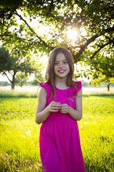 Portrait of little girl wearing vibrant pink dress smiling at camera with blade of grass in hands - LVF08139