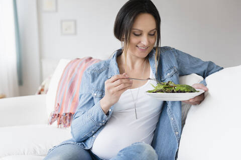 Schwangere kaukasische Frau isst Salat auf dem Sofa, lizenzfreies Stockfoto