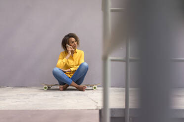 Porträt einer jungen Frau, die barfuß auf einem Skateboard sitzt und telefoniert - MOEF02330