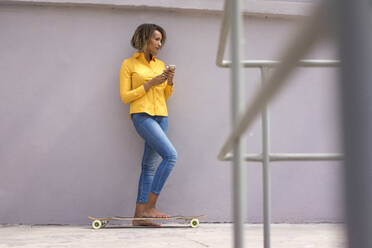 Young barefoot woman with smartphone and skateboard wearing yellow shirt and jeans - MOEF02331