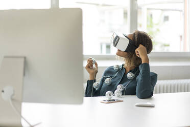 Young woman sitting at desk with chain of lights using Virtual Reality Glasses - MOEF02377