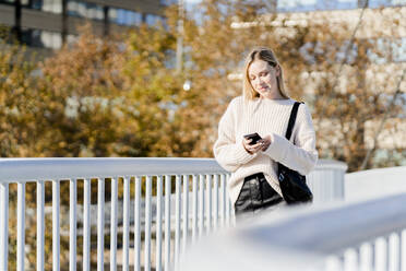 Porträt einer lächelnden jungen Frau, die im Herbst auf einer Fußgängerbrücke steht und auf ihr Mobiltelefon schaut - GIOF06588