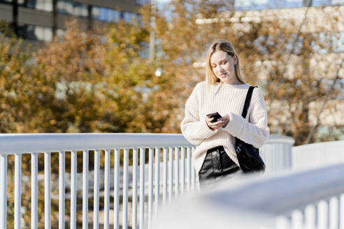 Porträt einer lächelnden jungen Frau, die im Herbst auf einer Fußgängerbrücke steht und auf ihr Mobiltelefon schaut - GIOF06588
