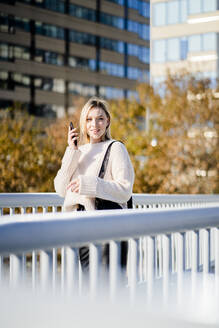 Porträt einer lächelnden jungen Frau mit Smartphone auf einer Fußgängerbrücke im Herbst - GIOF06589