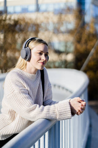 Porträt einer zufriedenen jungen Frau, die am Geländer einer Fußgängerbrücke lehnt und mit Kopfhörern Musik hört, lizenzfreies Stockfoto