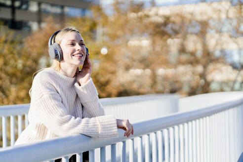 Porträt einer glücklichen jungen Frau, die am Geländer einer Fußgängerbrücke lehnt und mit Kopfhörern Musik hört - GIOF06591