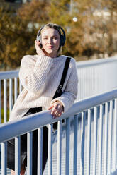 Portrait of content young woman leaning on railing of footbridge listening music with headphones - GIOF06592