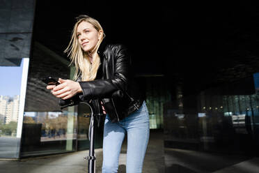 Porträt einer blonden jungen Frau mit Smartphone und Kick-Scooter in der Stadt - GIOF06610