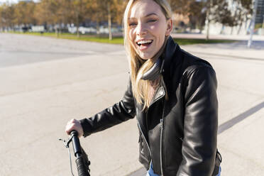 Portrait of laughing young woman with kick scooter wearing black leather jacket - GIOF06620