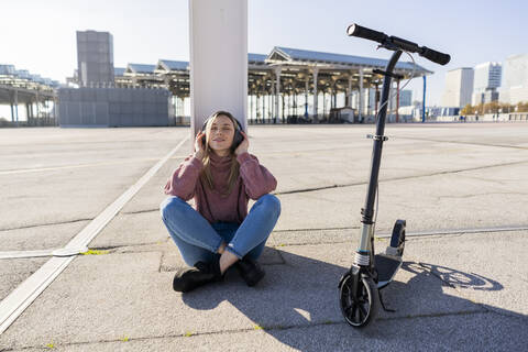 Junge Frau mit Kick-Scooter, die mit drahtlosen Kopfhörern Musik hört, Barcelona, Spanien, lizenzfreies Stockfoto