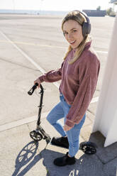 Portrait of smiling young woman with kick scooter and wireless headphones, Barcelona, Spain - GIOF06626