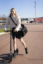 Lachende junge Frau mit Sporttasche auf einem Kick-Scooter - GIOF06638