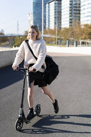 Lächelnde junge Frau mit Sporttasche auf einem Kick-Scooter, lizenzfreies Stockfoto