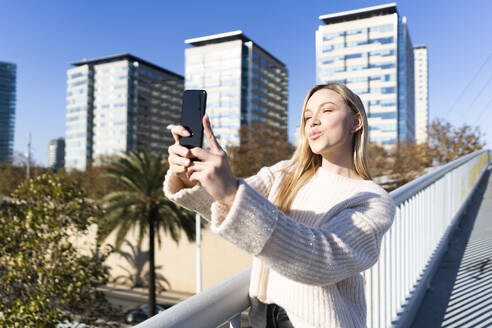 Porträt einer blonden jungen Frau auf einer Fußgängerbrücke, die ein Selfie mit ihrem Smartphone macht - GIOF06651