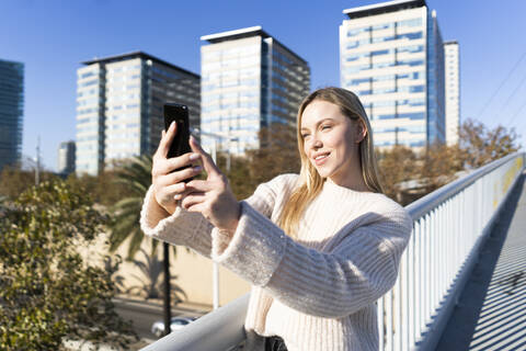 Porträt einer blonden jungen Frau auf einer Fußgängerbrücke, die ein Selfie mit ihrem Smartphone macht, lizenzfreies Stockfoto